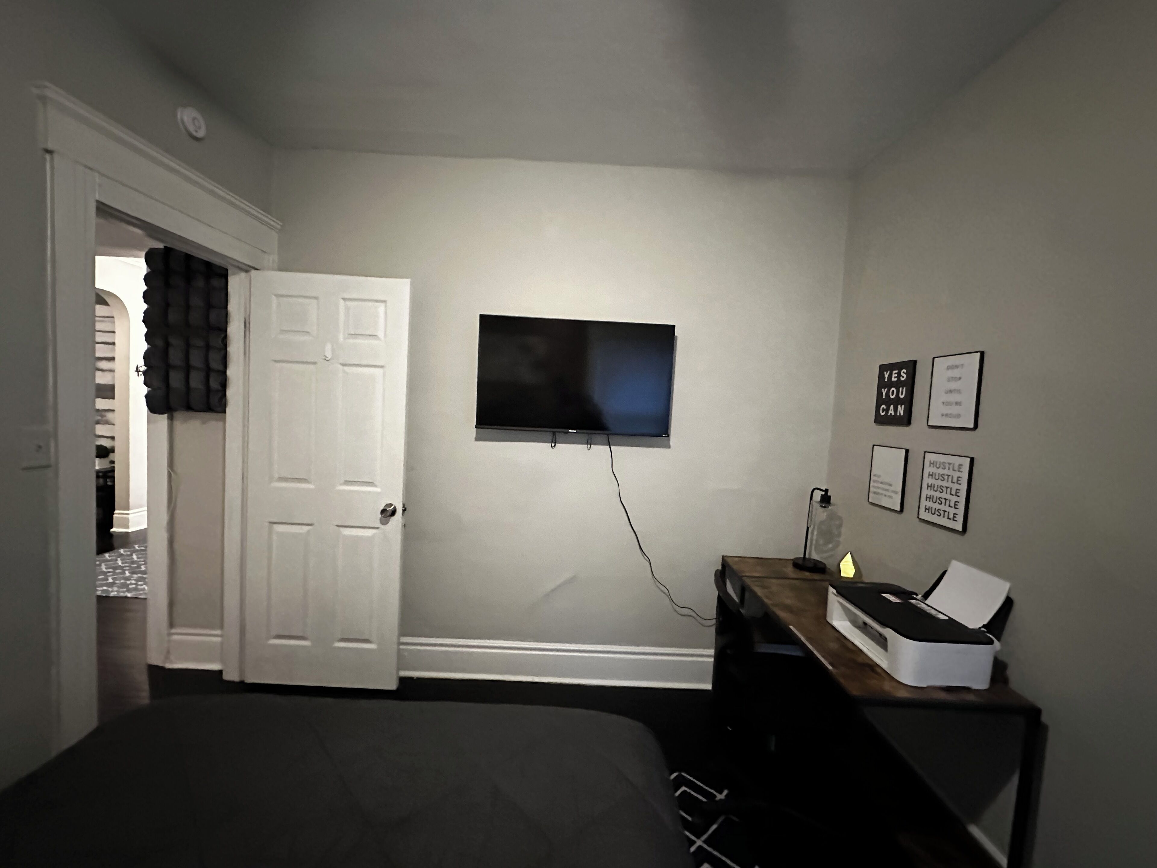 Un dormitorio con una cama, un escritorio con una impresora, un televisor montado en la pared y carteles motivacionales.