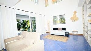 A spacious living room with a sofa, a coffee table, and a lamp. There is a window with blinds and a view of greenery outside.
