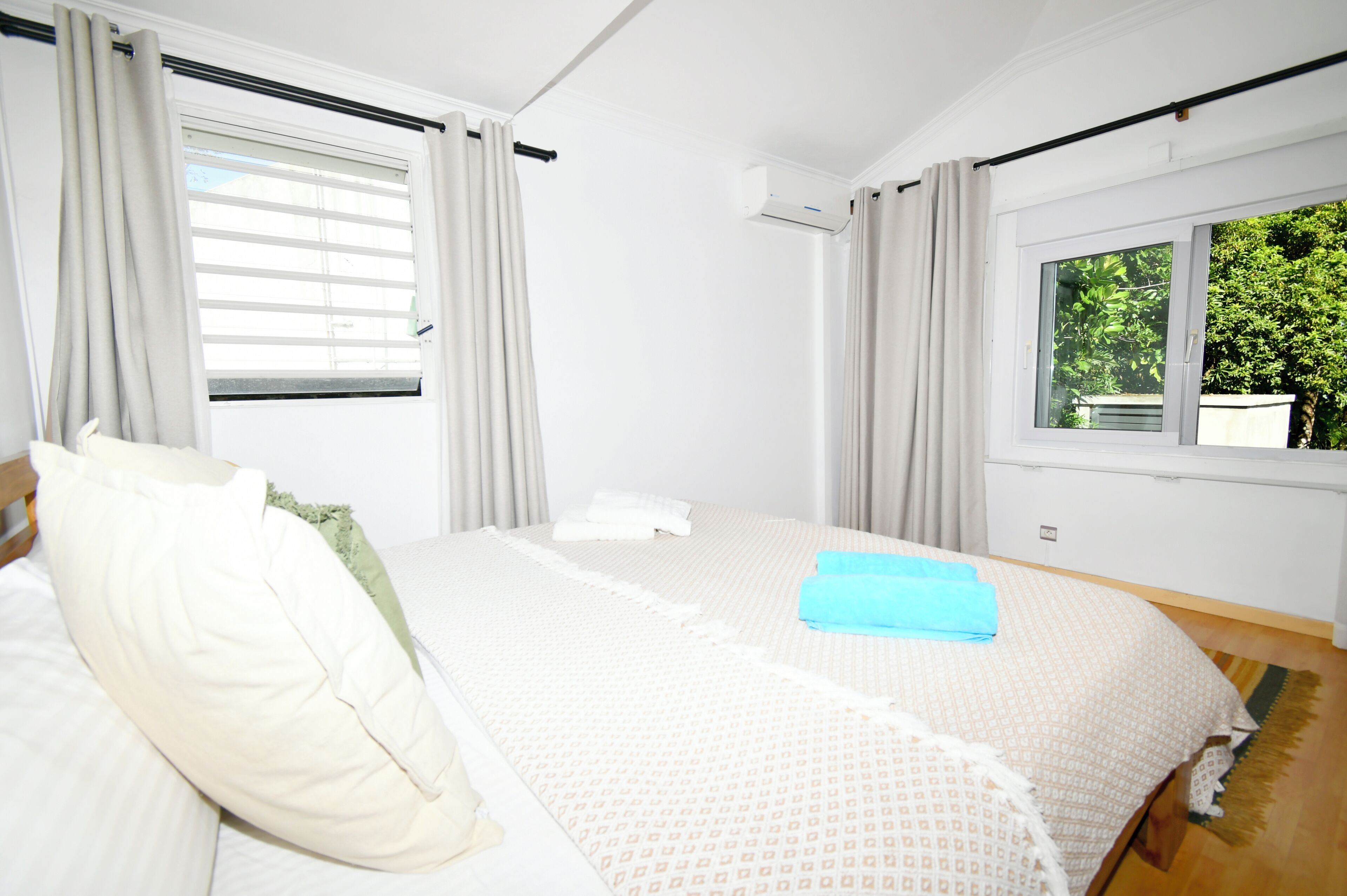 Un dormitorio con cama, cortinas, aire acondicionado y ventana con vistas a vegetación.