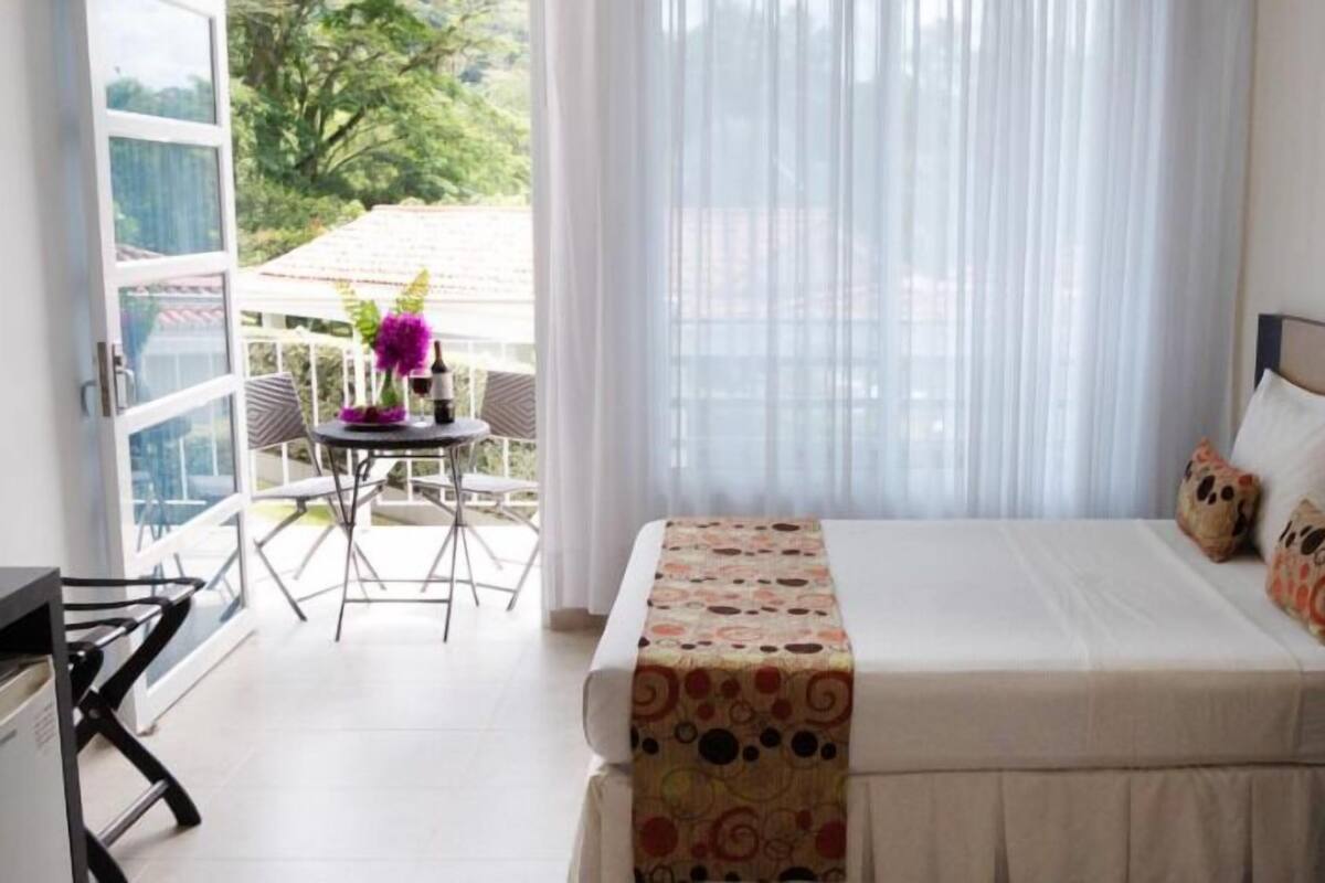 A hotel room with a bed, a small table with chairs, and a view of the outdoors.