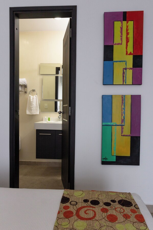 A hotel room with a bed, a bathroom with a sink and mirror, and two abstract paintings on the wall.