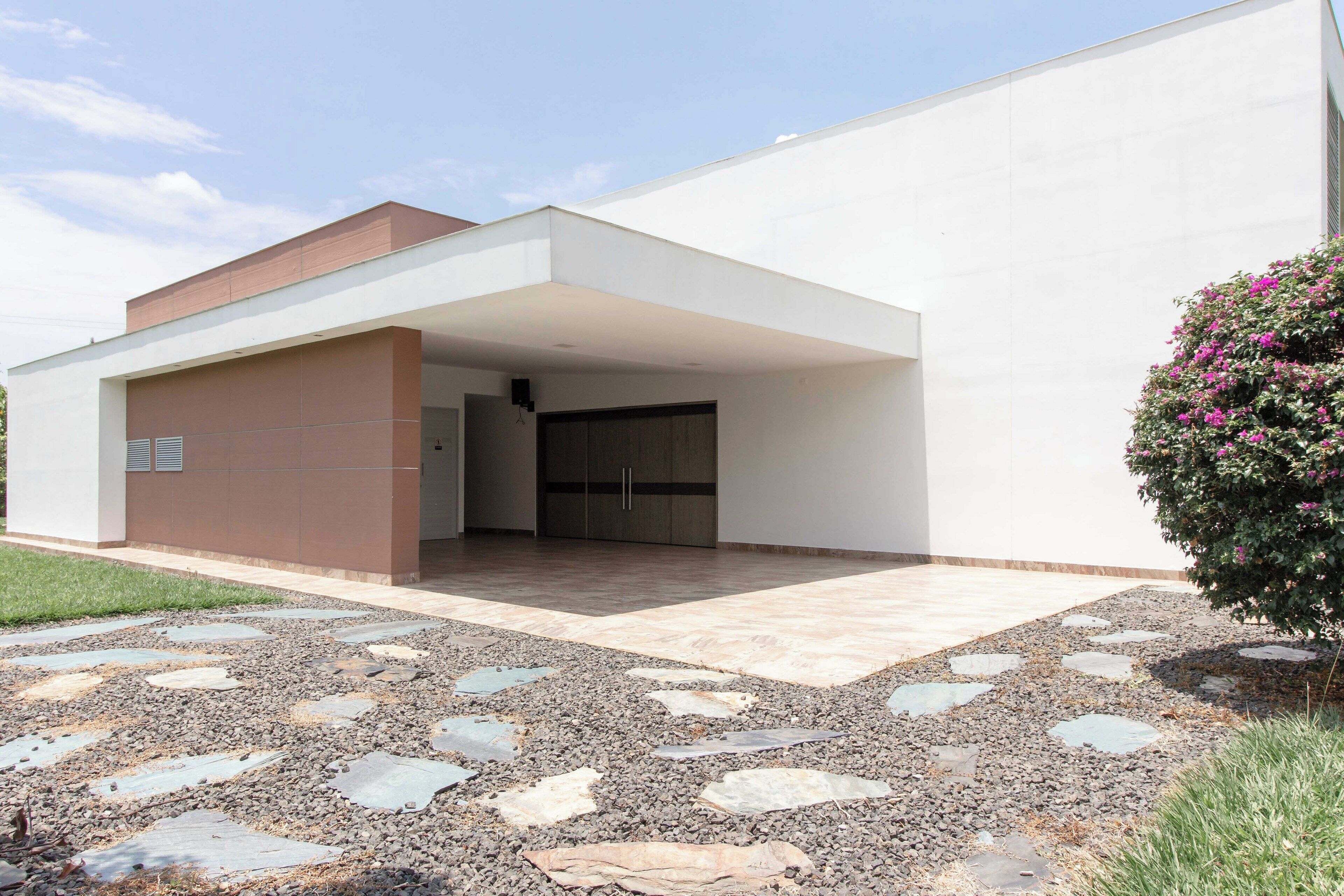 Un edificio moderno con un acceso cubierto, un camino de grava y un arbusto pequeño con flores rosadas.
