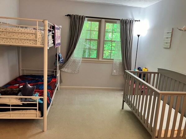 A child's room with bunk beds, a crib, a window with curtains, and a wall-mounted shelf.