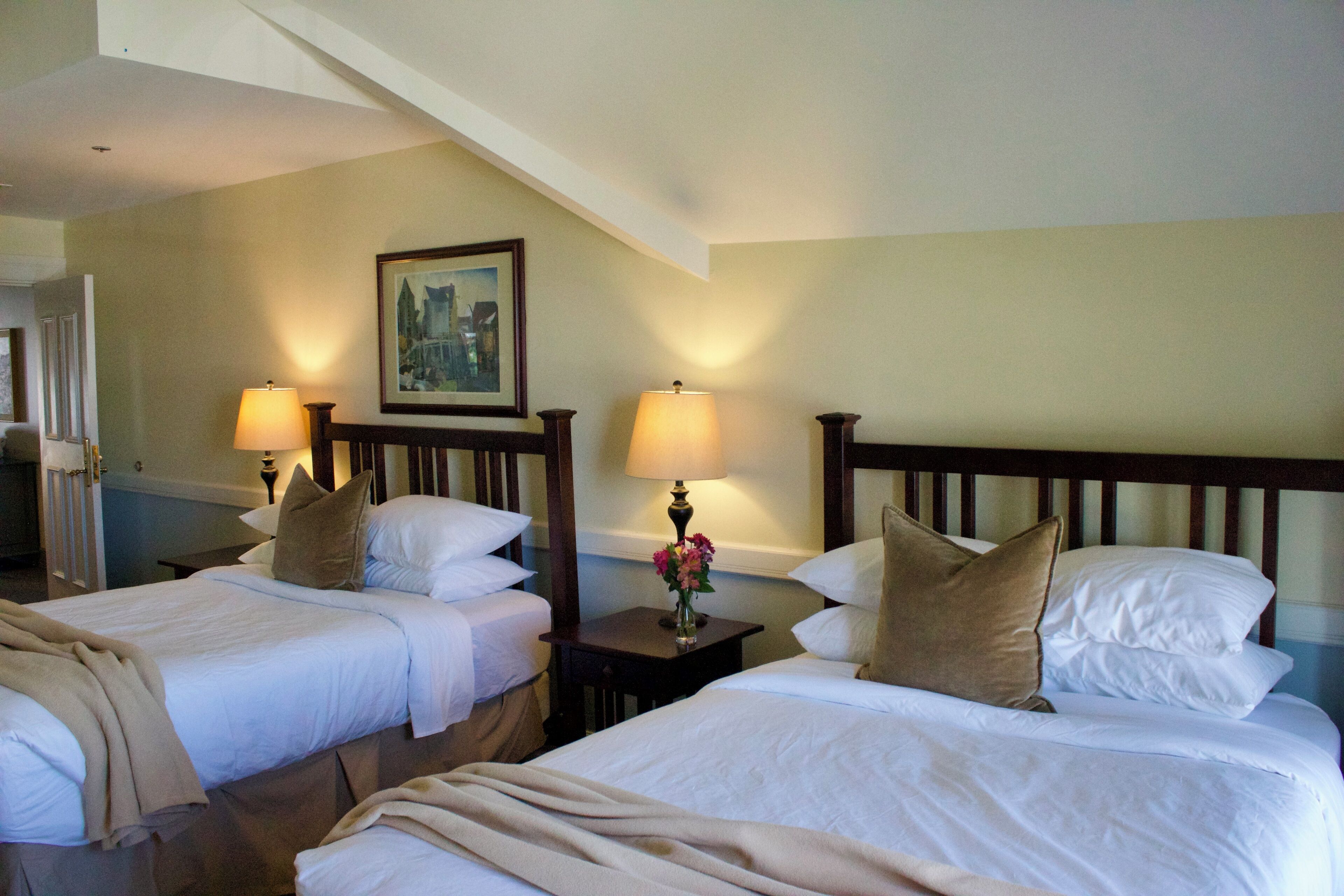A hotel room with two beds, each with white linens and brown pillows. There are two bedside lamps and a framed picture on the wall.