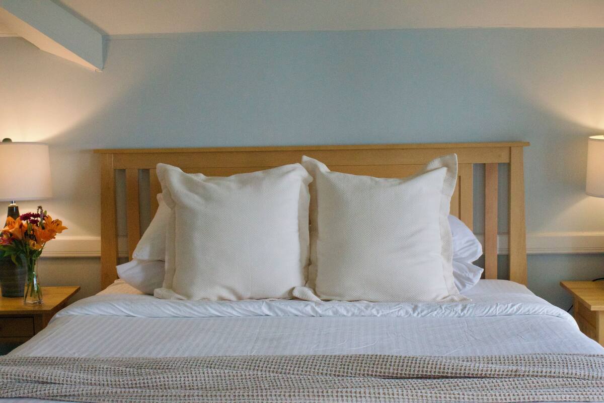 A neatly made bed with white linens and two pillows, a wooden headboard, and a nightstand with a vase of flowers.