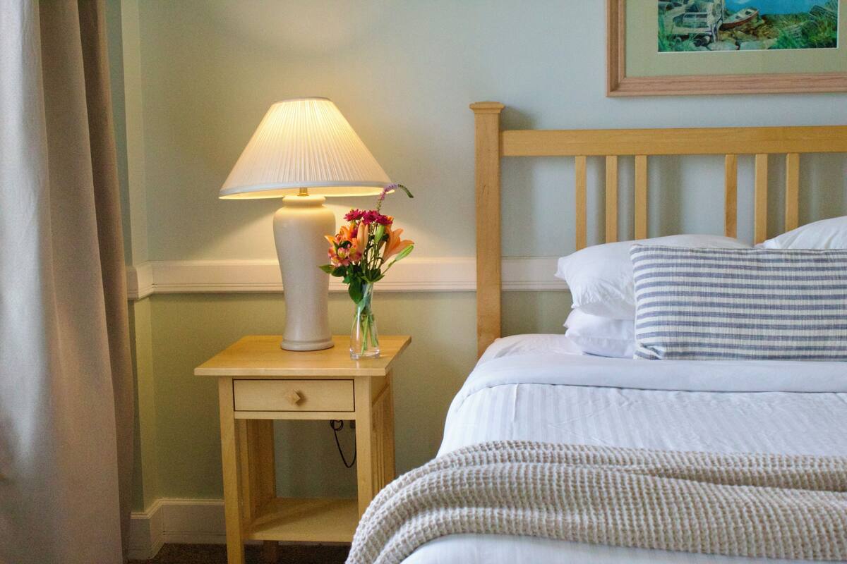 A neatly made bed with white linens, a bedside lamp, and a vase with flowers on a wooden nightstand.