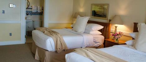 A hotel room with two beds, a bathroom, and a nightstand with a lamp and flowers.