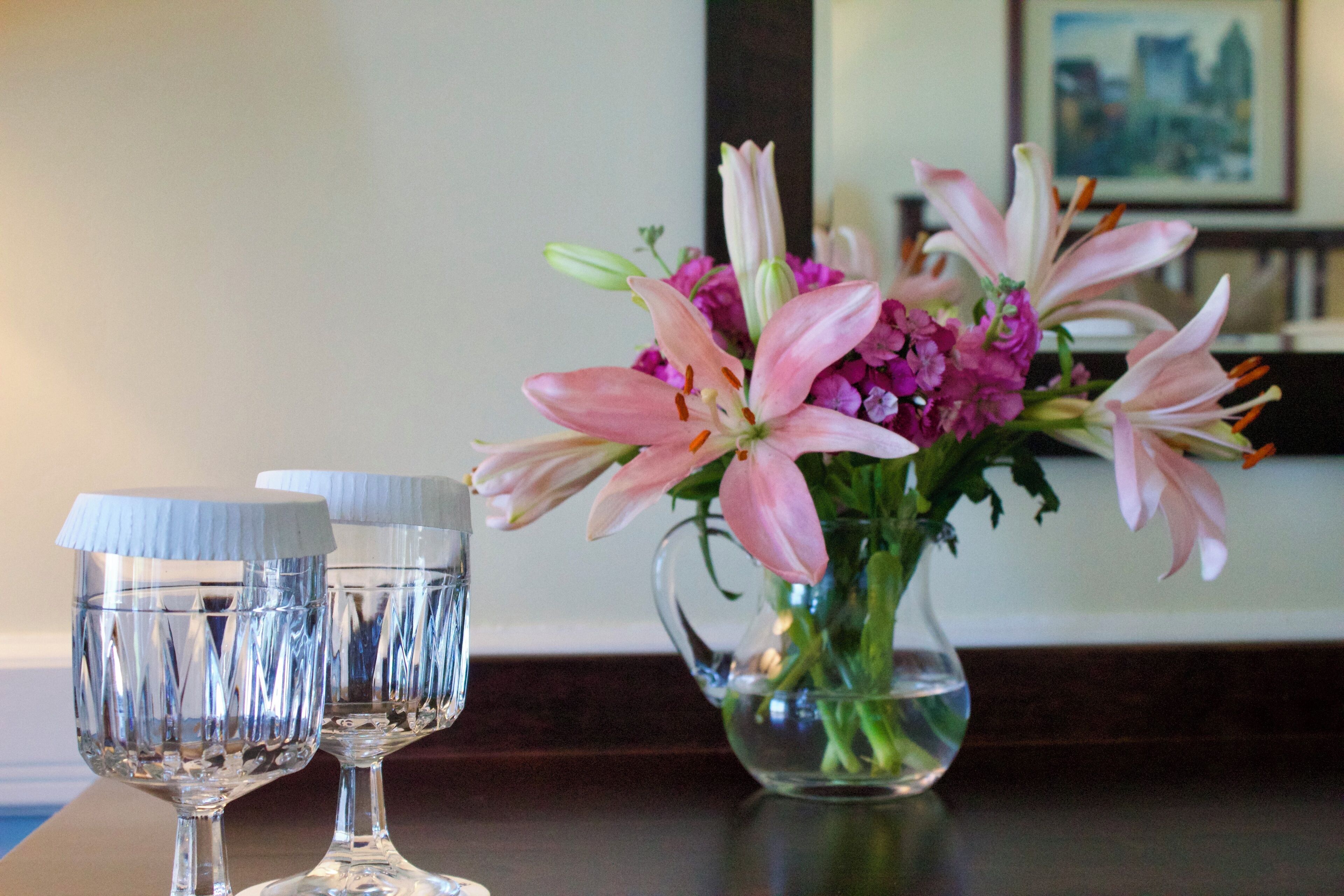 Two clear glass goblets on a wooden table next to a vase with pink and purple flowers.