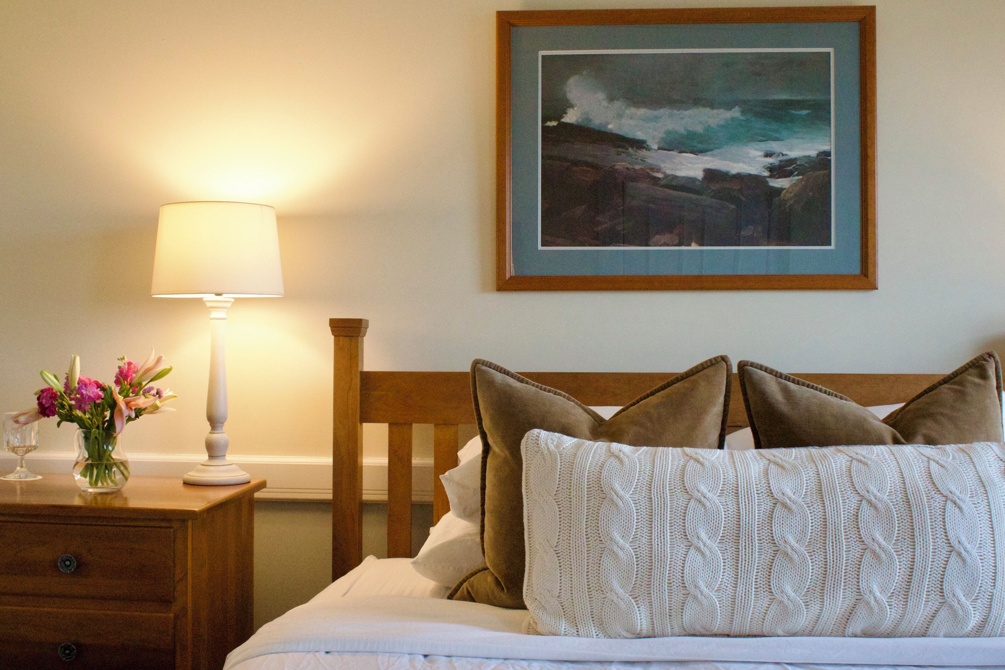 A neatly made bed with a white comforter and brown pillows, a bedside table with a lamp and a vase of flowers, and a framed painting of a coastal scene on the wall.