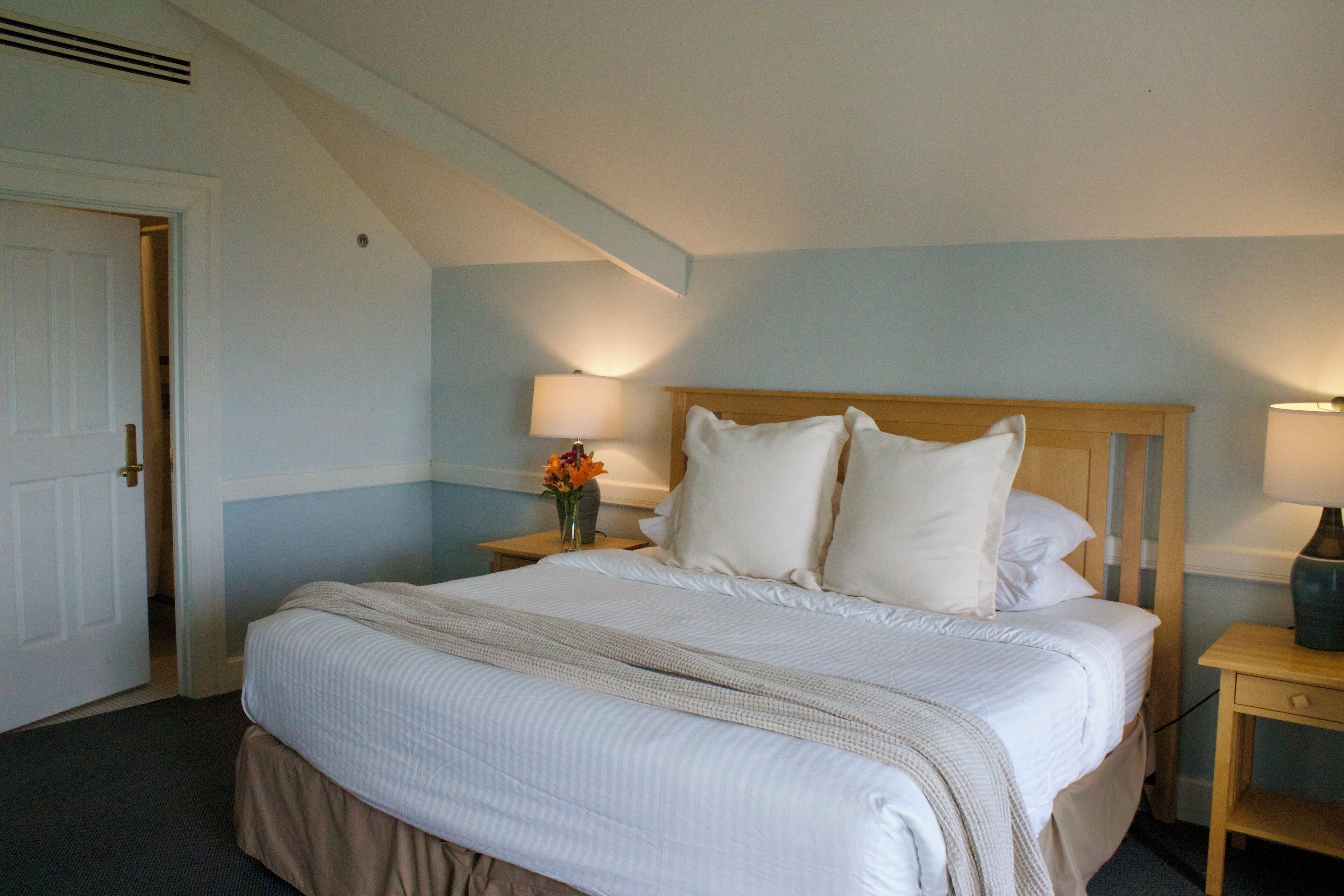 A neatly made bed with white linens and a beige throw blanket in a room with light blue walls and a sloped ceiling.