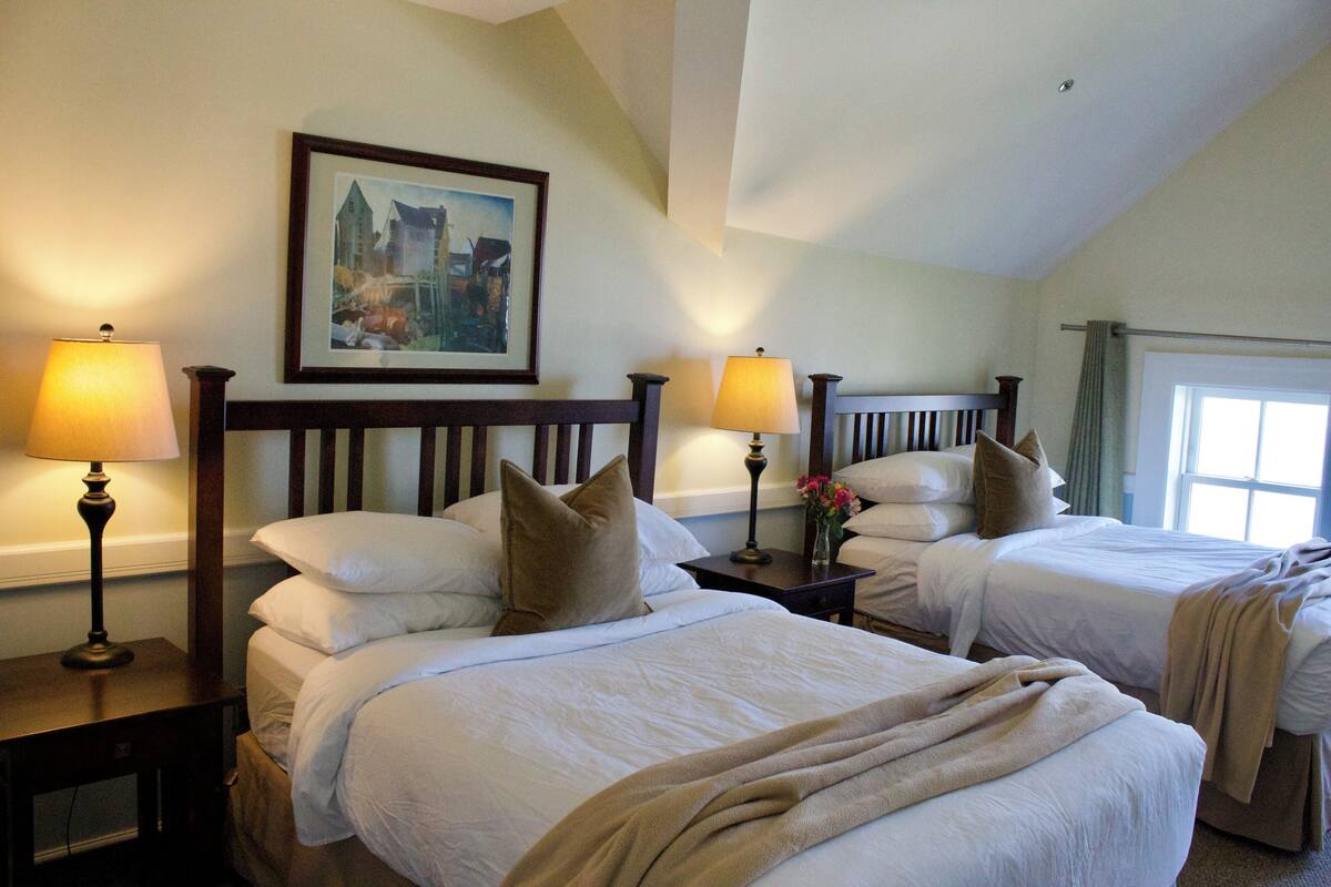 A hotel room with two beds, each with a wooden headboard, white bedding, and a beige throw blanket. There are two bedside tables with lamps and a framed picture on the wall.
