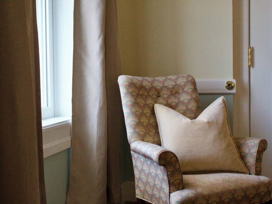 A corner chair with a patterned fabric and a cushion, positioned near a window with curtains.