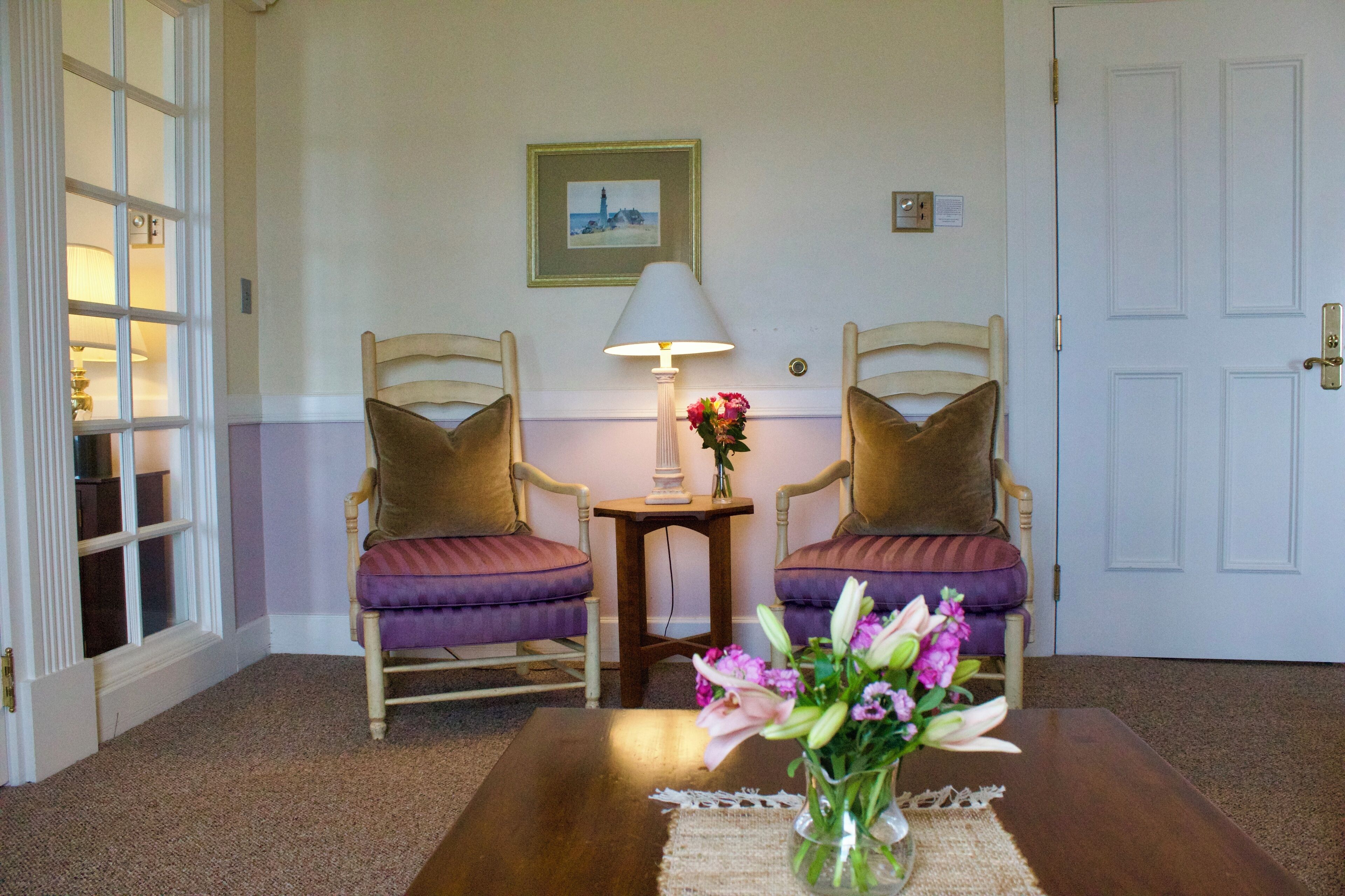 A room with two wooden chairs with purple cushions, a small table with a vase of flowers, a lamp, and a framed picture on the wall.
