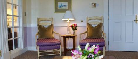 A room with two wooden chairs with purple cushions, a small table with a vase of flowers, a lamp, and a framed picture on the wall.