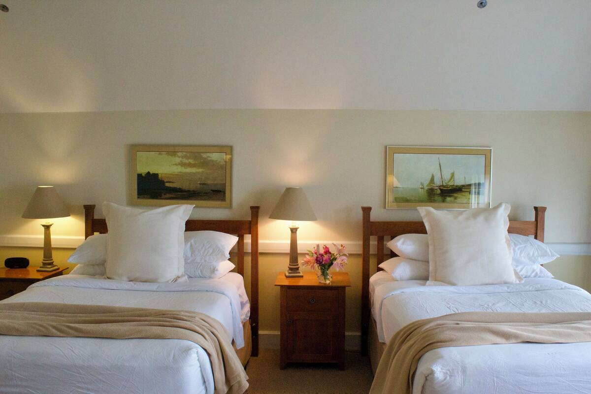 A hotel room with two beds, each with white bedding and a wooden headboard. There are two bedside tables with lamps and a small vase of flowers on one table.
