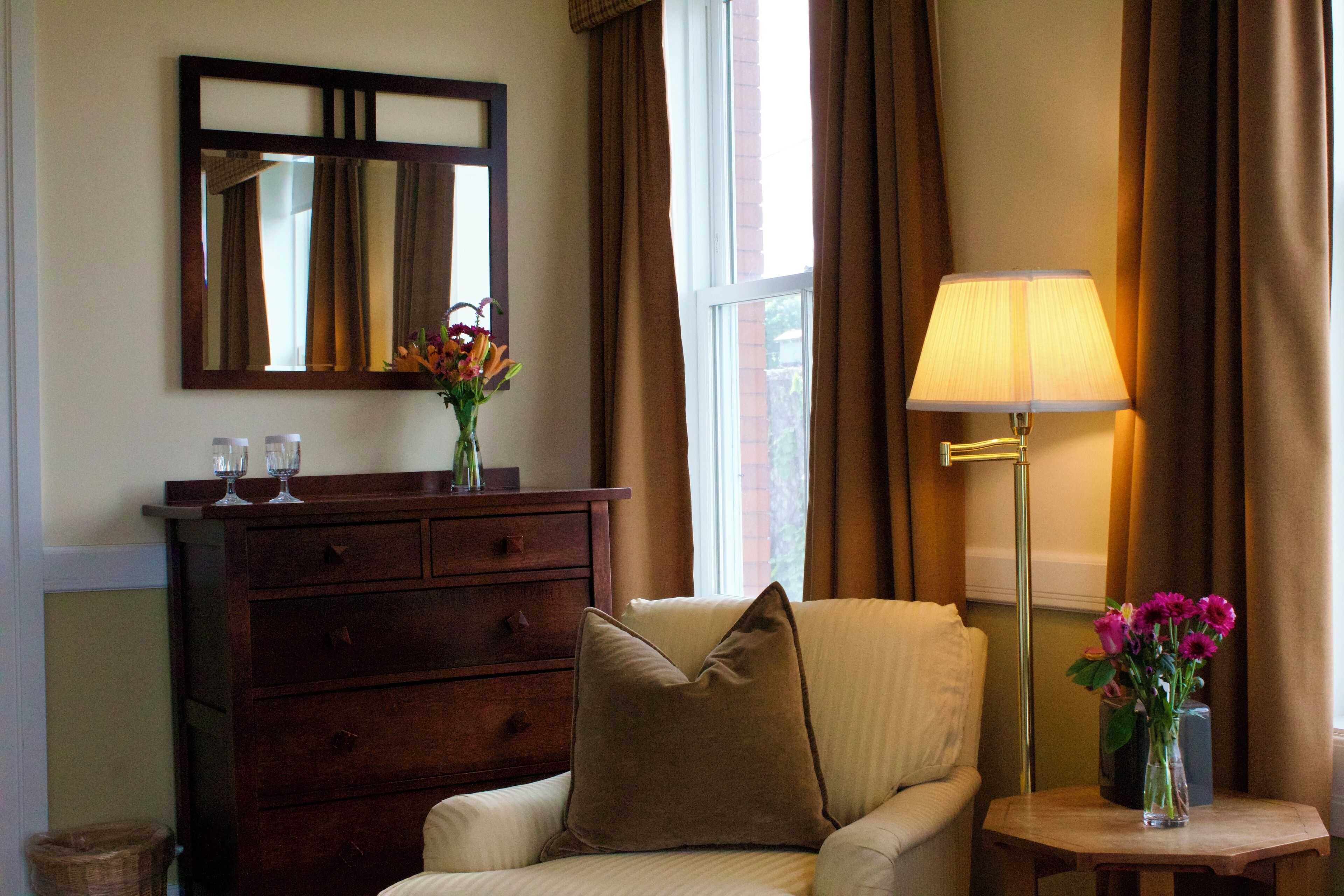 A hotel room with a wooden dresser, a cream-colored armchair, a small table with a lamp and flowers, and a large mirror on the wall.
