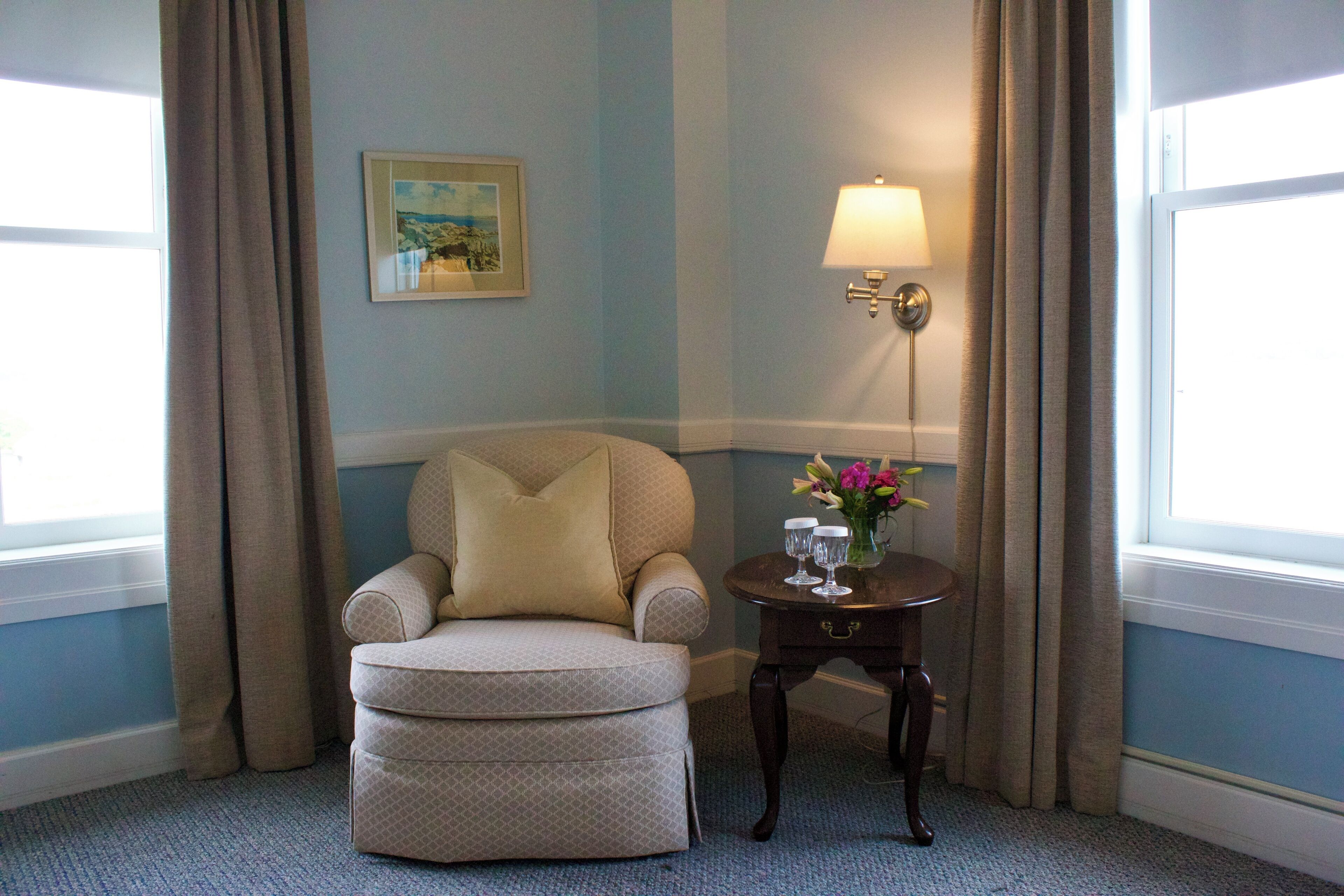 Ein Zimmer mit einem beigefarbenen Sessel, einem kleinen Tisch mit einer Lampe und einem Blumenstrauß sowie einem gerahmten Bild an der Wand.