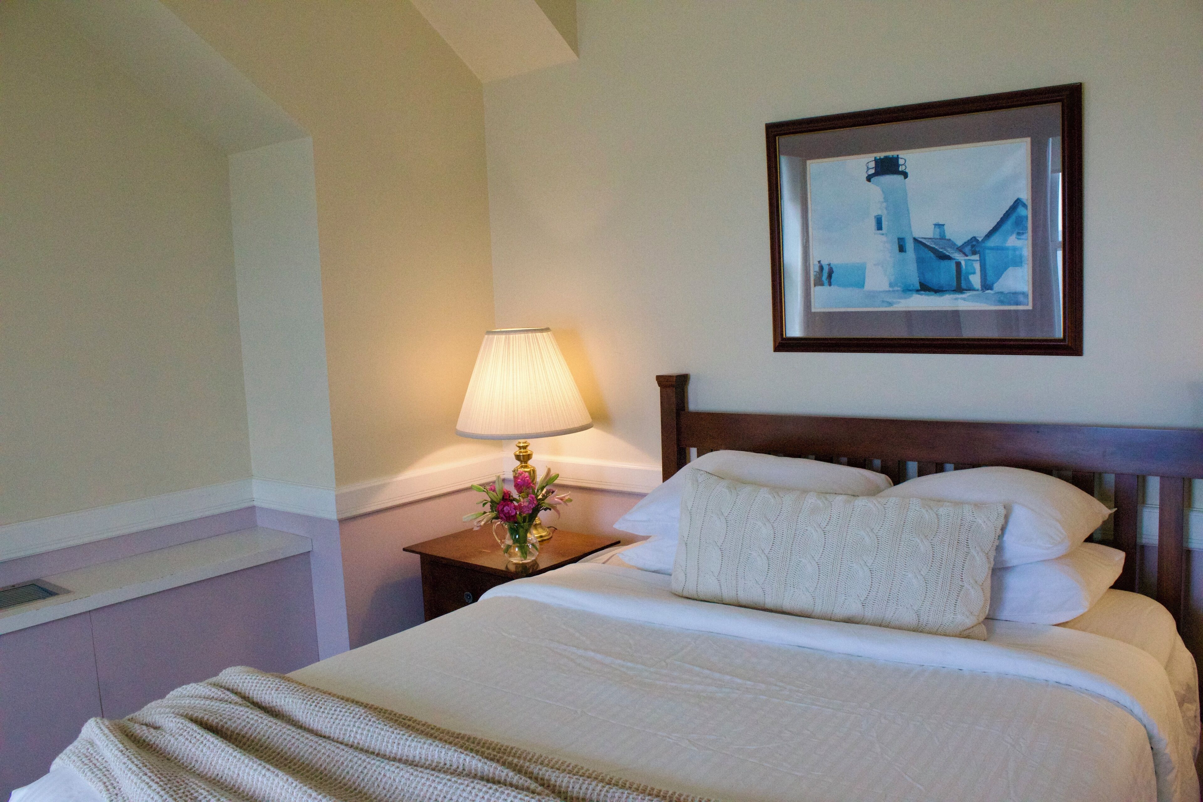 A neatly made bed with a white comforter and pillows, a bedside lamp with a floral arrangement, and a framed picture of a lighthouse on the wall.
