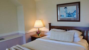 A neatly made bed with a white comforter and pillows, a bedside lamp with a floral arrangement, and a framed picture of a lighthouse on the wall.