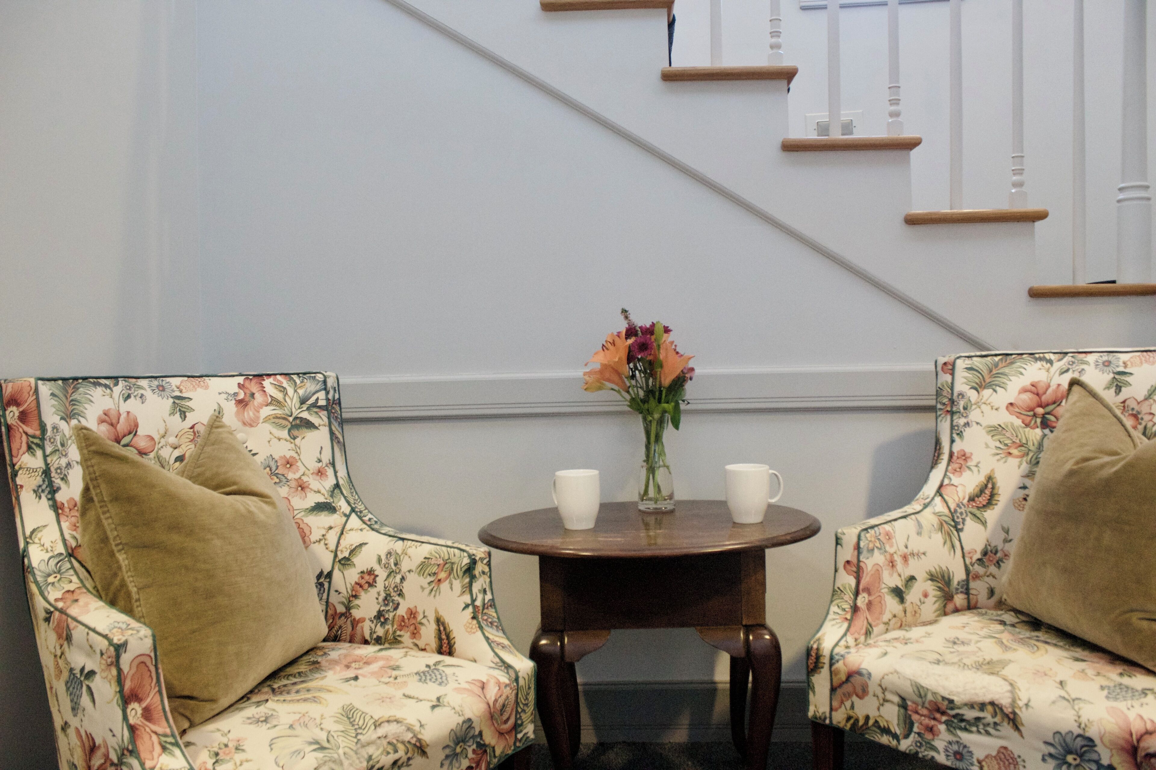 Two floral-patterned chairs with cushions, a small round table with a vase and two white cups, and a staircase with wooden steps and white railings.