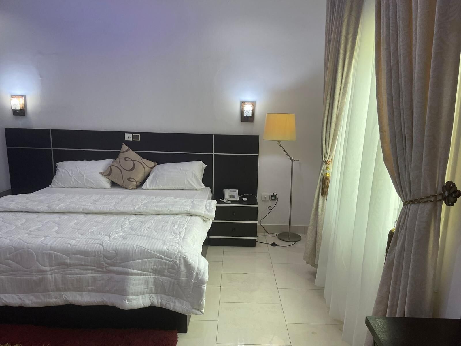 Una cama bien hecha con ropa blanca, una mesita de noche con lámpara y un teléfono en la pared.
