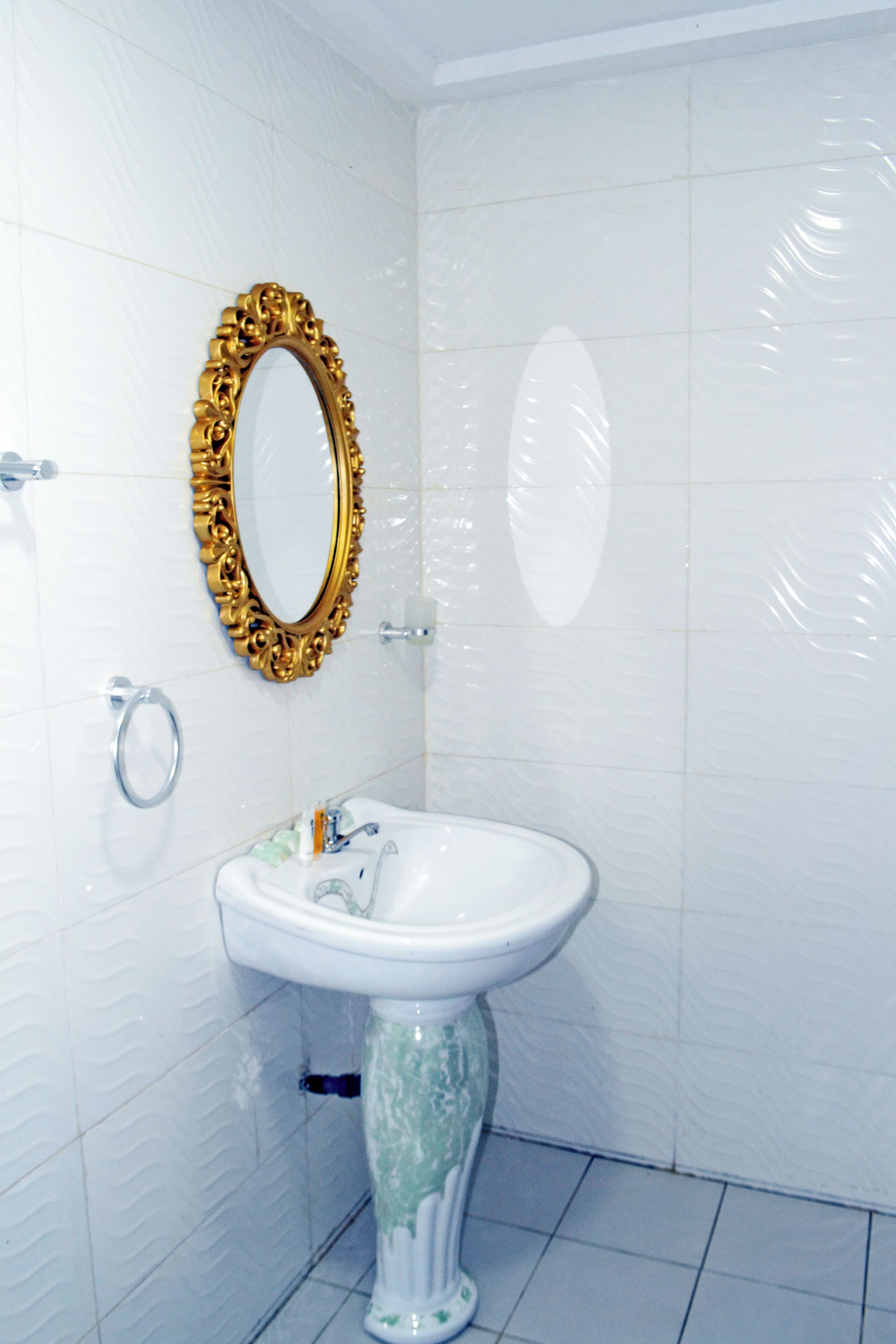 Un baño con paredes de azulejos blancos, un lavamanos pedestal blanco, un espejo con marco dorado y un inodoro con un dispensador de papel higiénico plateado.
