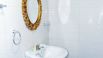 Un baño con paredes de azulejos blancos, un lavamanos pedestal blanco, un espejo con marco dorado y un inodoro con un dispensador de papel higiénico plateado.