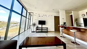 A modern living room with a large window, a flat-screen TV, a wooden coffee table, and a kitchen area with a bar. - Adorable 1-bedroom apartment in tranquil Queretaro perfect for relaxing (QUERETARO)