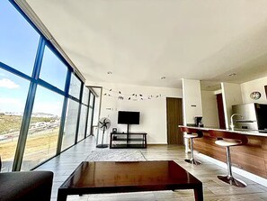 Ein modernes Wohnzimmer mit großem Fenster, einem Flachbildfernseher, einem Couchtisch aus Holz und einer Küchenzeile mit Bar.