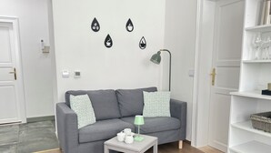A modern living room with a grey sofa, patterned pillows, a small side table with a white mug and a green lamp, and a wall-mounted coffee machine.