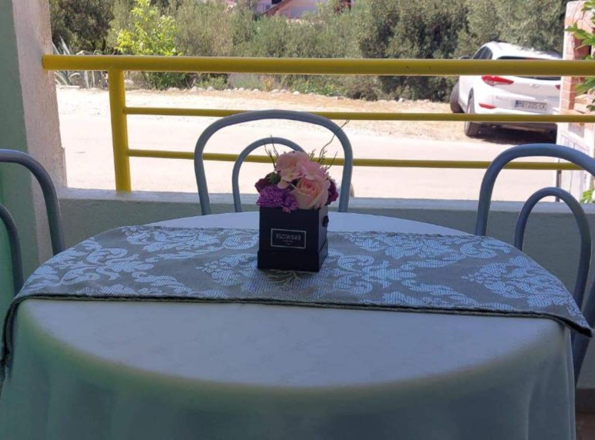 A table with a patterned tablecloth, a black vase with pink flowers, and metal chairs.