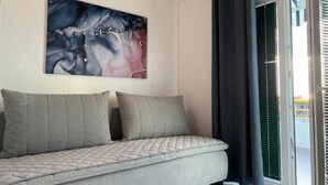 A modern living room with a grey sofa, a large abstract painting, and a sliding glass door with green shutters. - Apartments Natali (Gradac)