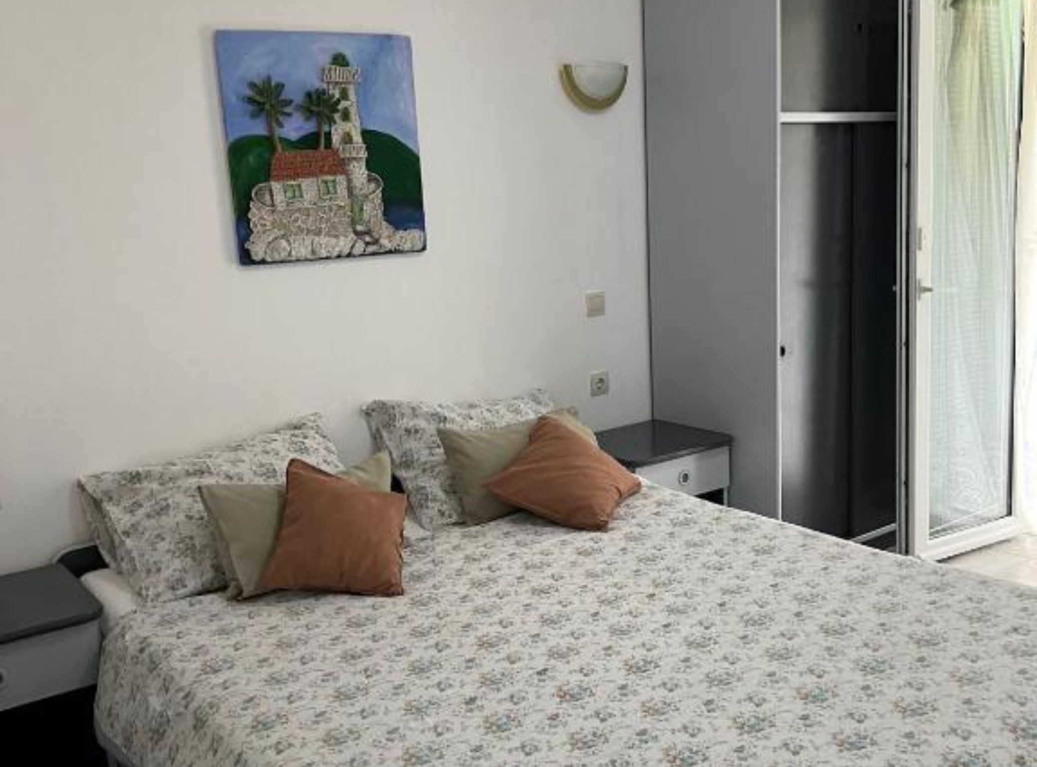 A bedroom with a bed, two pillows, a nightstand, a painting of a lighthouse, and a sliding door.
