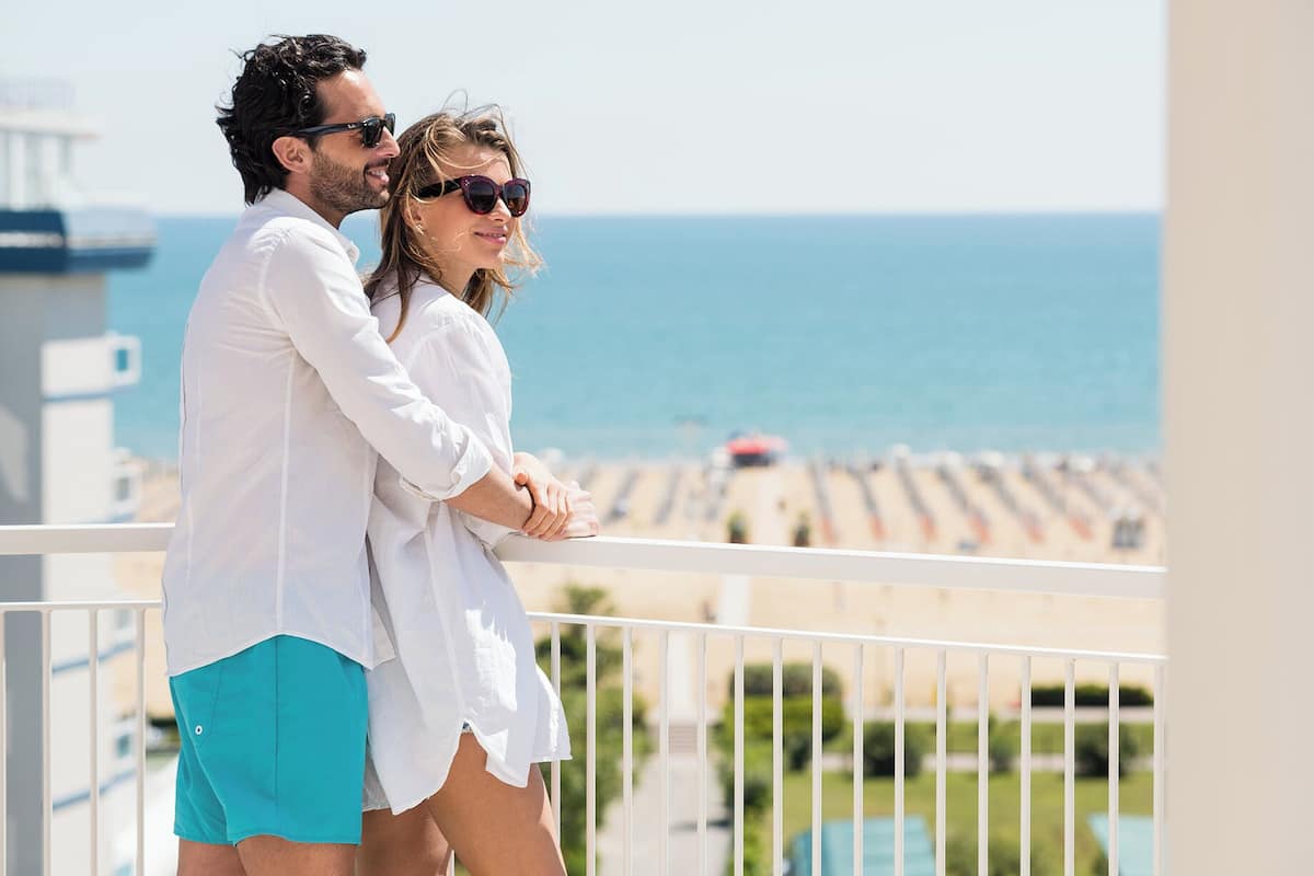Una pareja en un balcón con vistas a una playa, una piscina y un edificio al fondo.