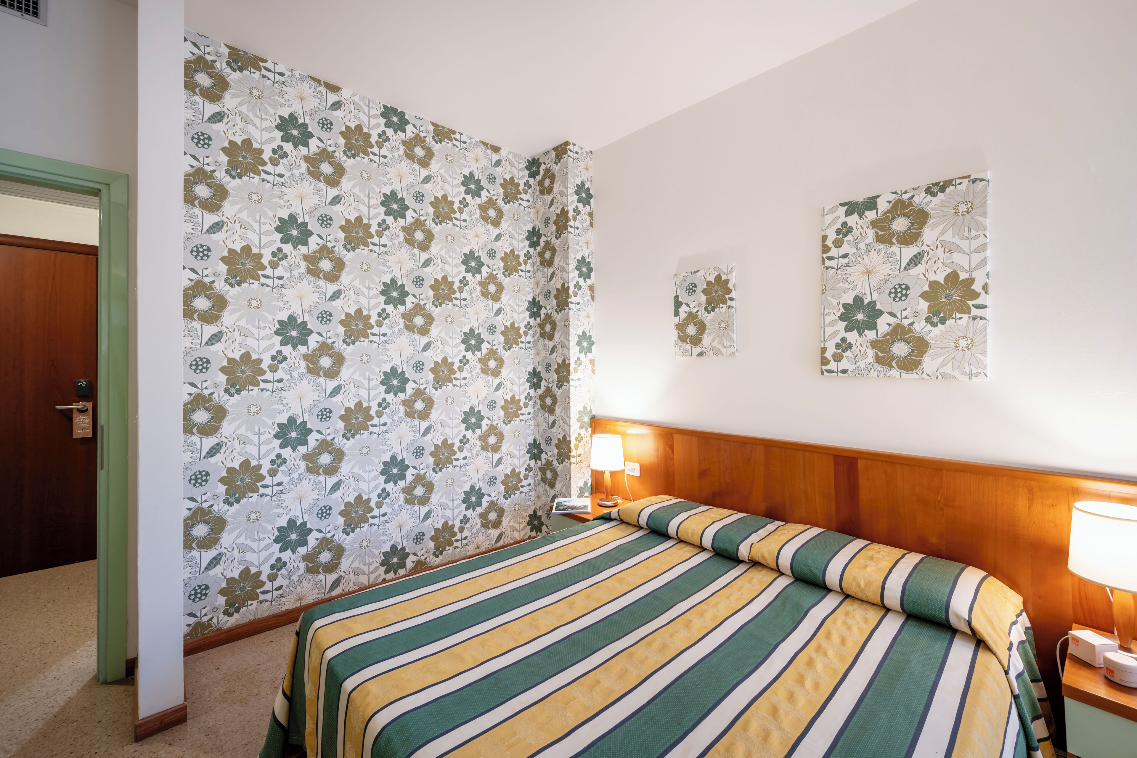 A hotel room with a bed, a nightstand, a lamp, floral wallpaper, and a door.