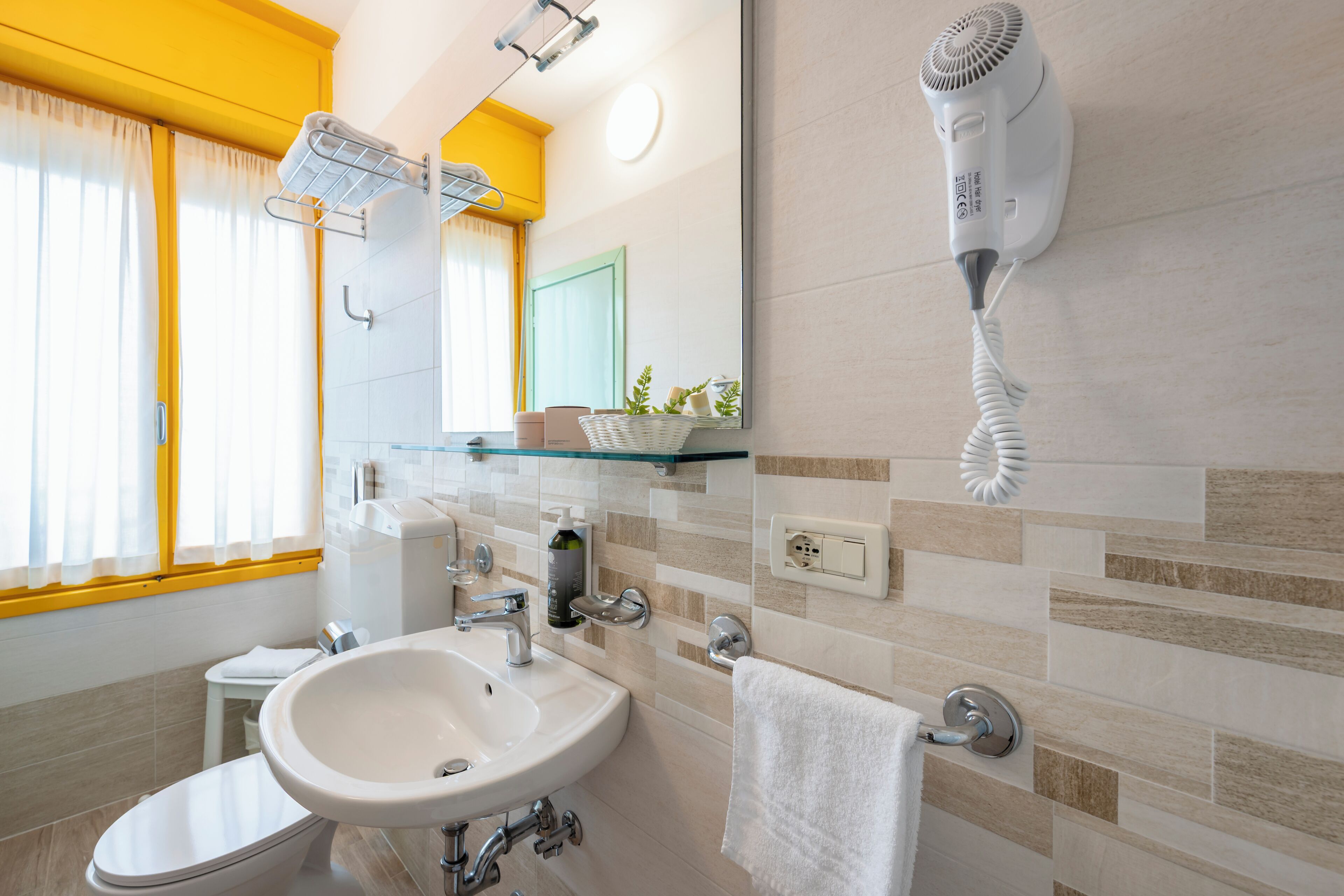 Un baño moderno con un lavabo blanco, un secador de pelo montado en la pared y un estante de vidrio con artículos de aseo.