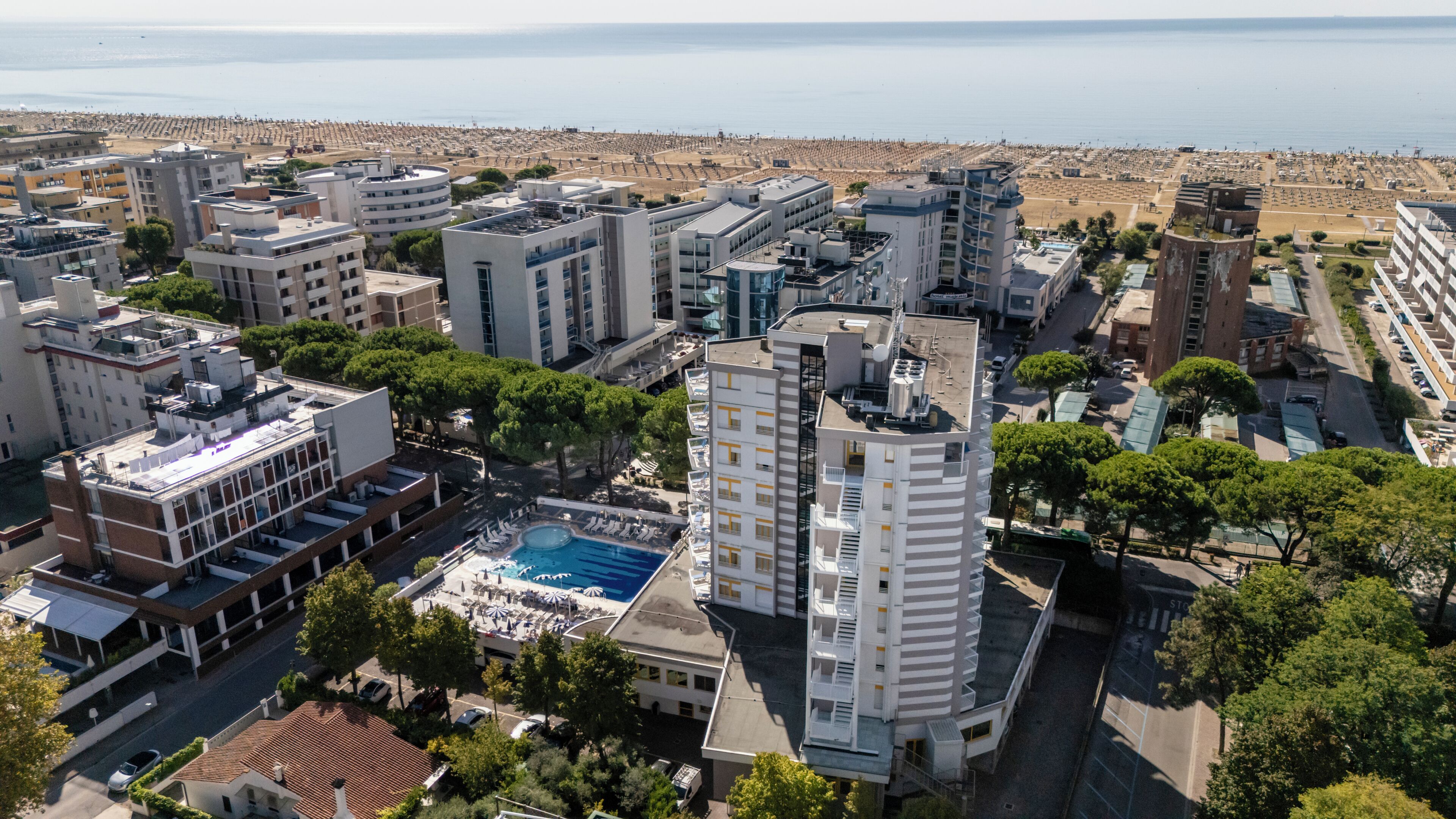 Ein Hochhaus-Hotel mit Pool auf dem Dach, umgeben von anderen Gebäuden und einem Strand im Hintergrund.