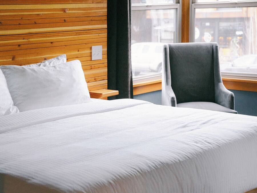 A hotel room with a large bed, a wooden headboard, a grey armchair, and a window with a view of the street.