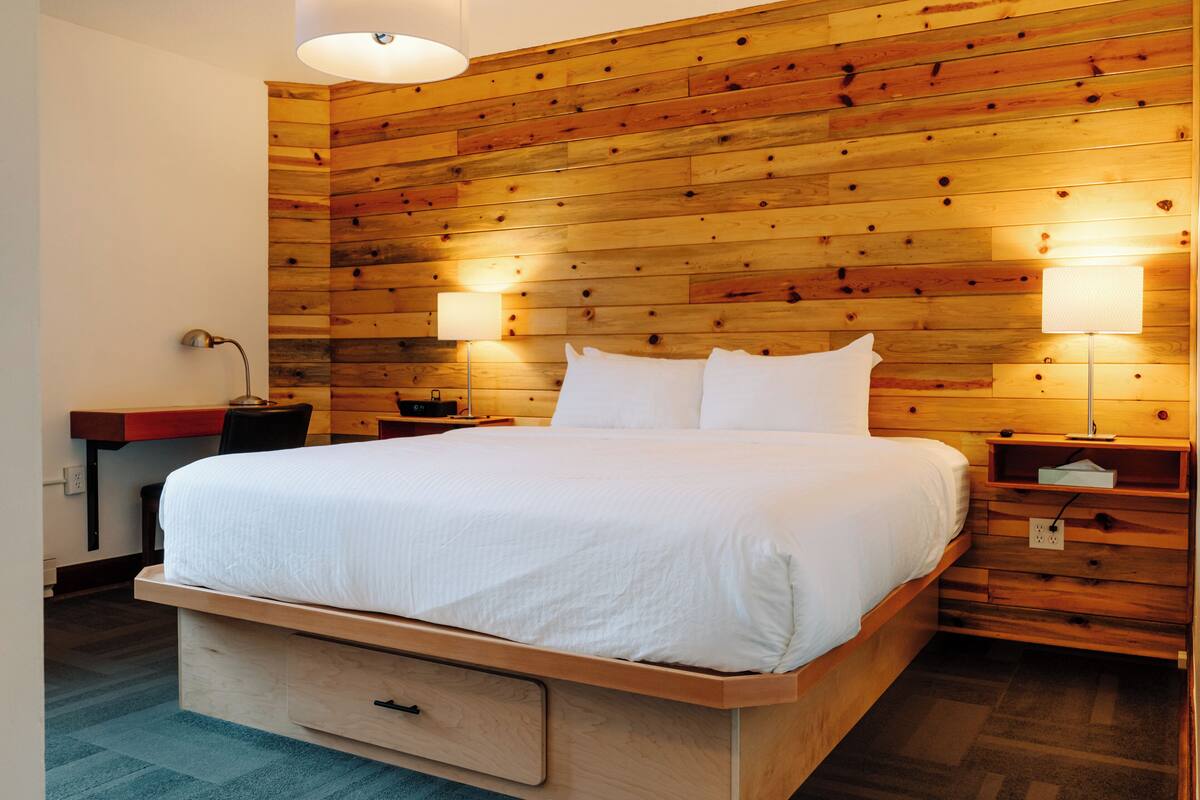 A neatly made bed in a room with wooden panel walls, a desk with a lamp, and a nightstand with a lamp.