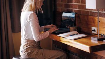 A person working at a wooden desk with a laptop, notebook, and pen. There are two black stools and a white lamp.