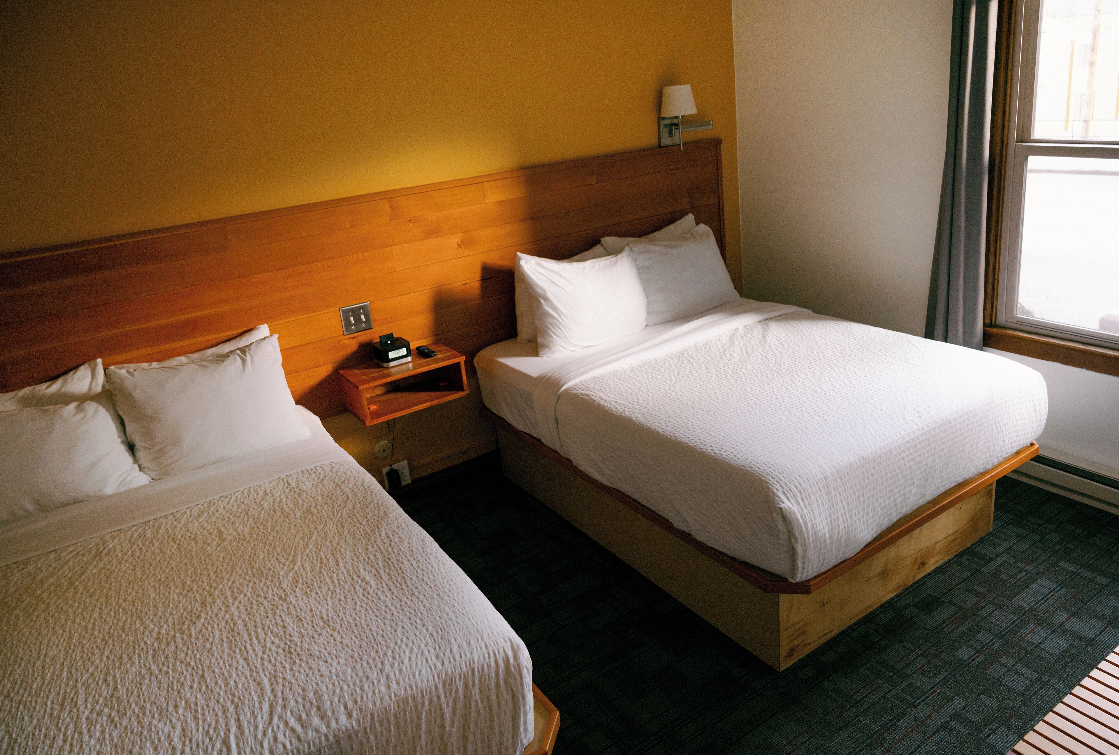 A hotel room with two single beds, wooden headboards, a nightstand with a lamp, and a window with a view.