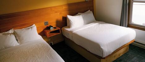 A hotel room with two single beds, wooden headboards, a nightstand with a lamp, and a window with a view.