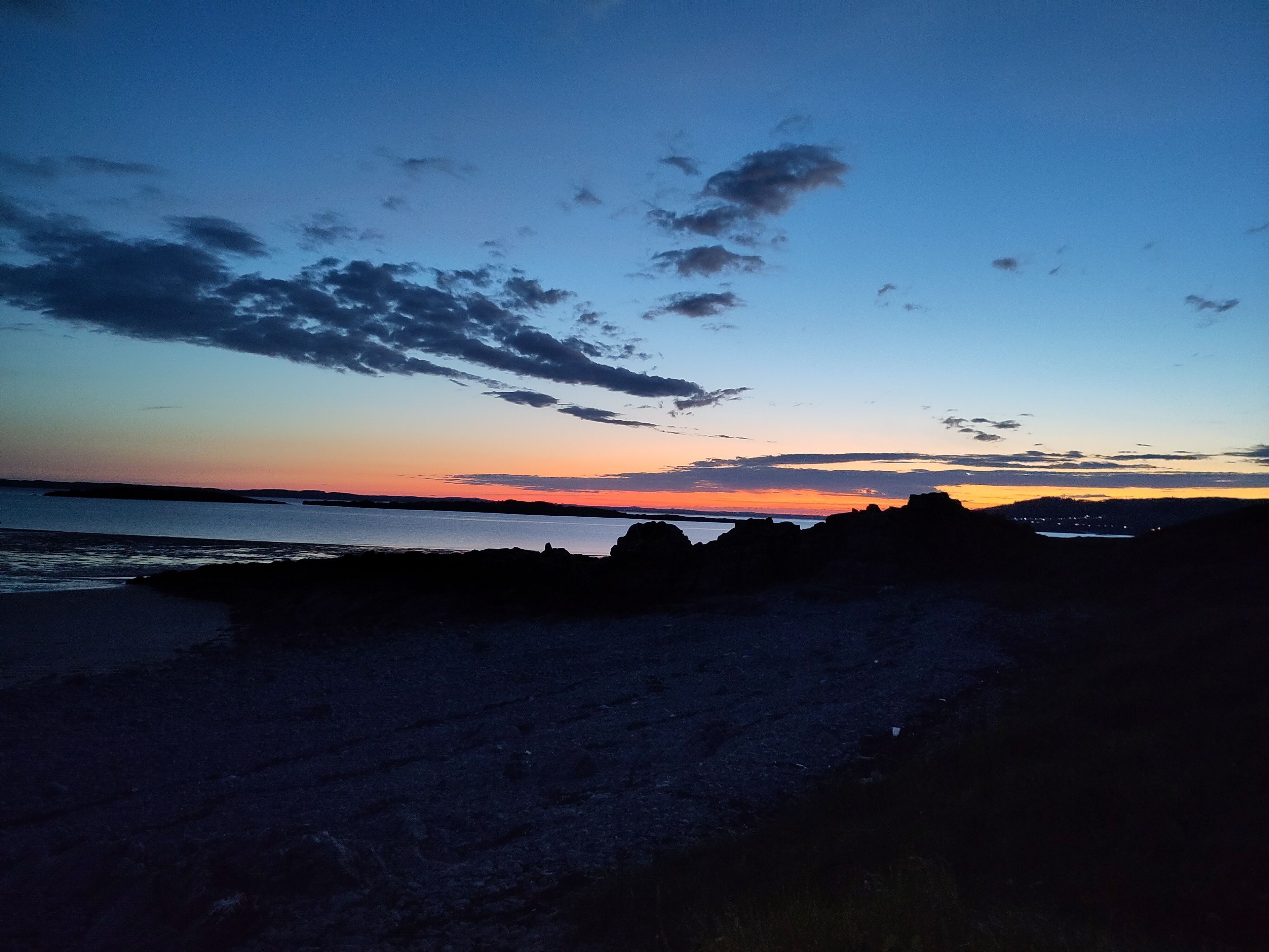 Un paysage côtier avec un coucher de soleil, une côte rocheuse et des lumières scintillantes au loin.