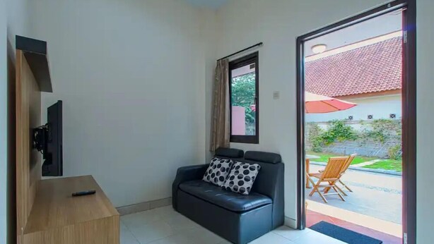 A small room with a black leather sofa, a wooden side table, a TV, and a sliding glass door leading to an outdoor area with a red umbrella and a chair.