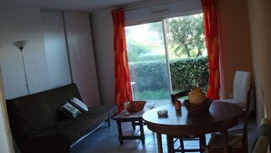 A living room with a sofa, a dining table, and chairs. There is a window with curtains, and a view of greenery outside. - T2 rez de Jardin 50m2 Bord de mer (Frontignan plage)