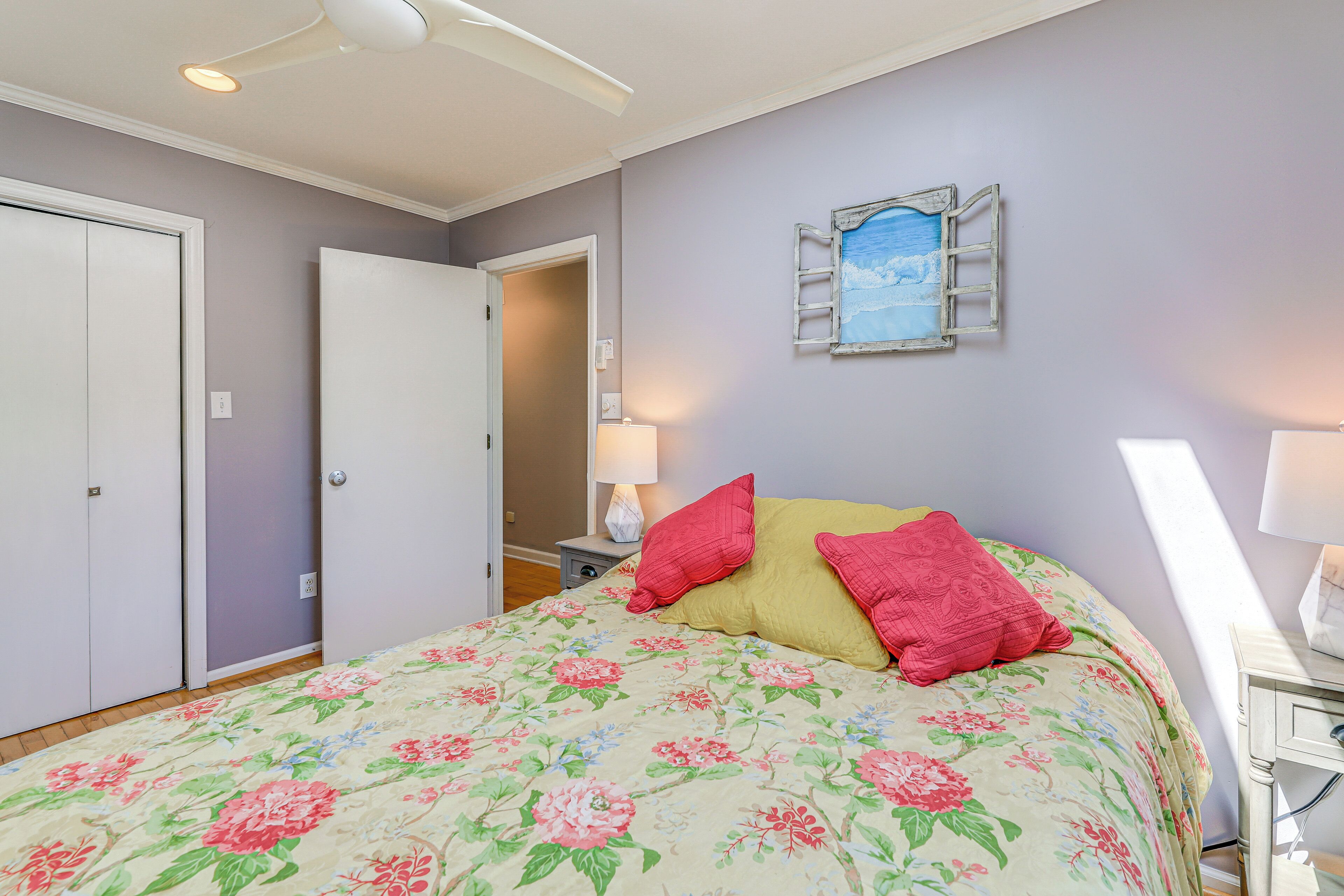 Une chambre à coucher avec une couvre-lit à motifs floraux, une table de chevet avec une lampe et une vue sur l’extérieur par une fenêtre.