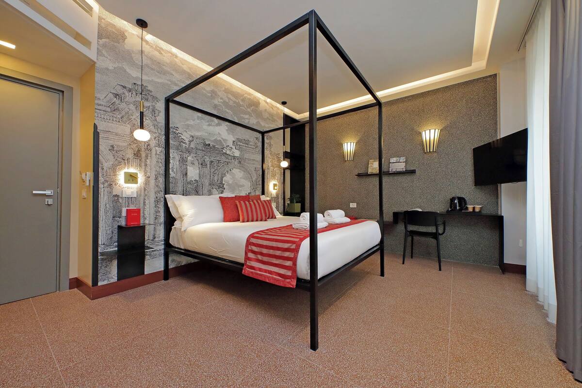 A modern hotel room with a four-poster bed, a desk with a chair, a flat-screen TV, and a decorative wall with a light fixture.