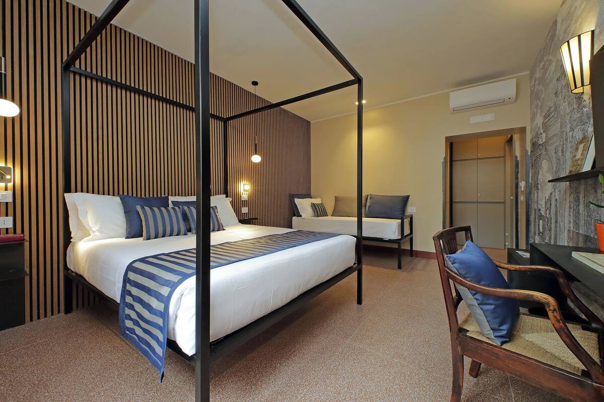 A four-poster bed with a striped blanket, a wooden chair with a blue cushion, a desk with a lamp, and a stone wall.