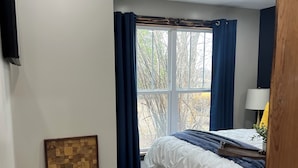 A bedroom with a bed, a window with curtains, a wall-mounted air conditioner, a potted plant, and a decorative wooden panel. - Cozy Cabin - Fire pit, Upscale Area, Near I44 & HWY 65 (Springfield)