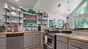 A modern kitchen with grey cabinets, a stainless steel stove, and a dishwasher. There are shelves with dishes and a window providing natural light. - Cozy Whispering Hideaway- Indulge in serenity and unwind in nature. (Ponchatoula)